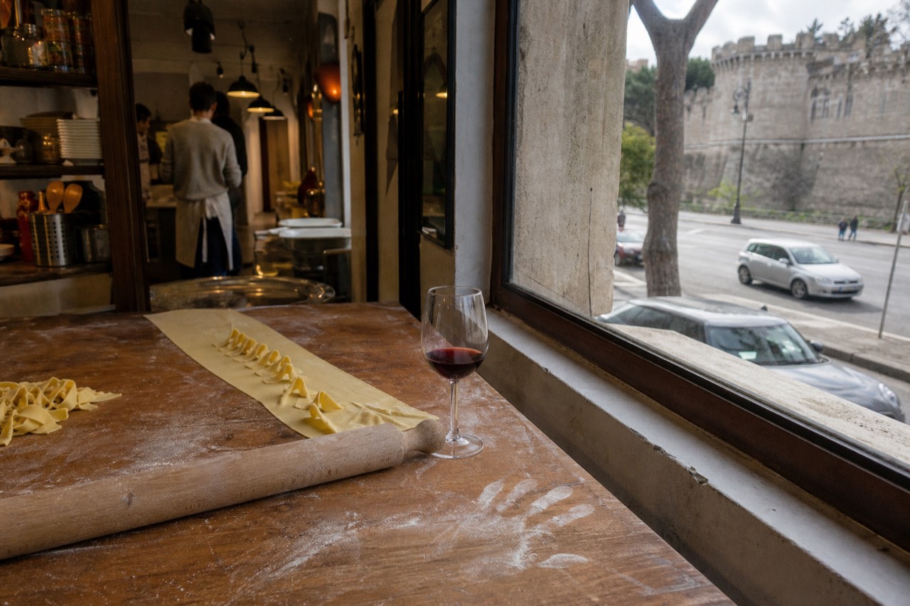 A rustic kitchen counter with fresh pasta dough, a rolling pin and a glass of red wine
