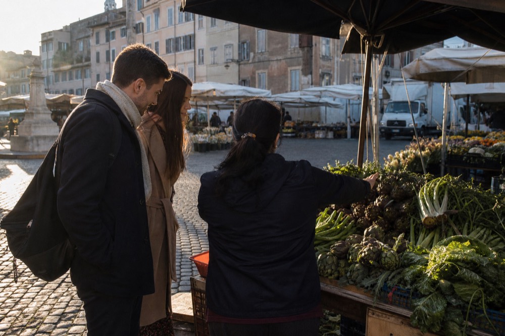 A guide speaking to a small group at a market stall piled with artichokes and leafy greens