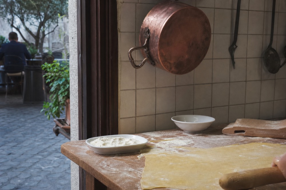 Fresh pasta dough being rolled by hand on a floured wooden board in a cooking school kitchen