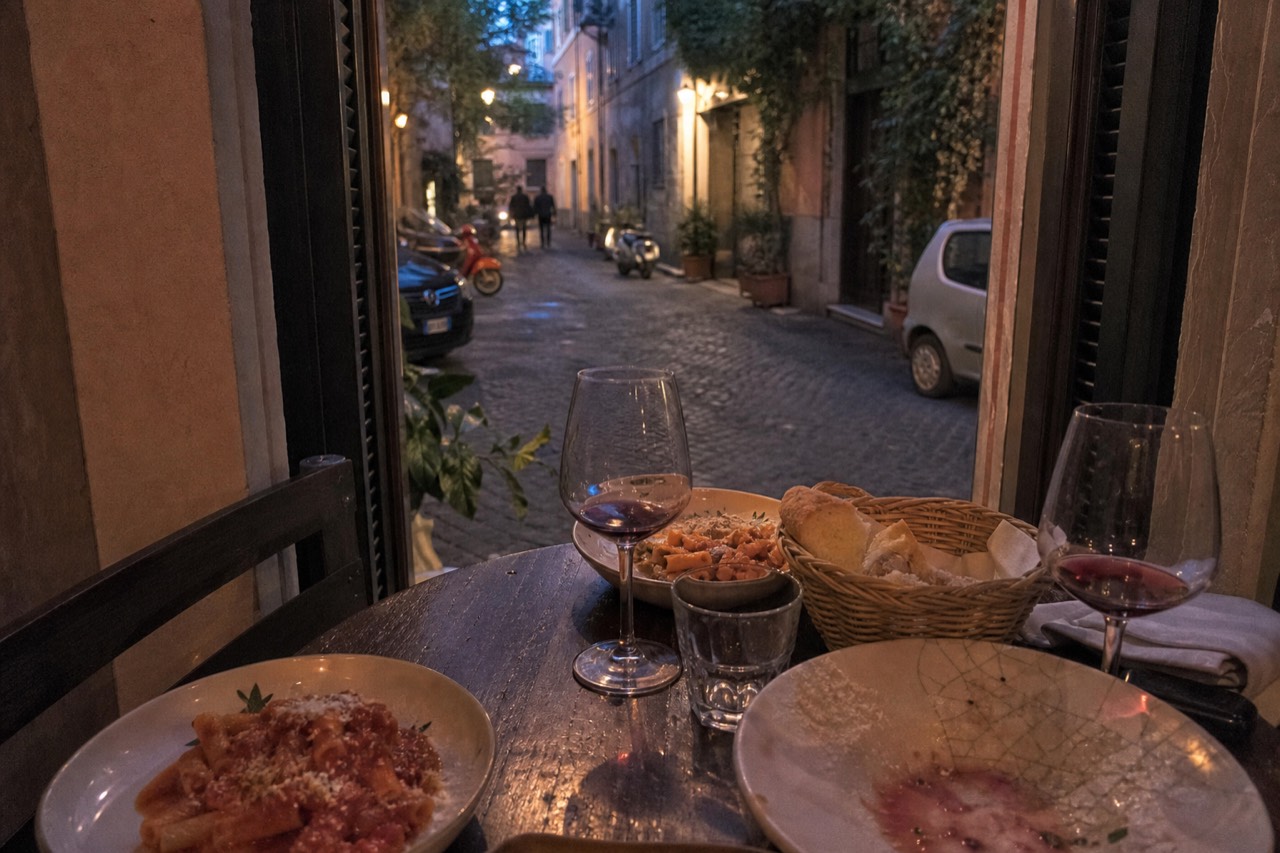A wooden table in a Roman trattoria holding plates of pasta, glasses of red wine and a basket of bread