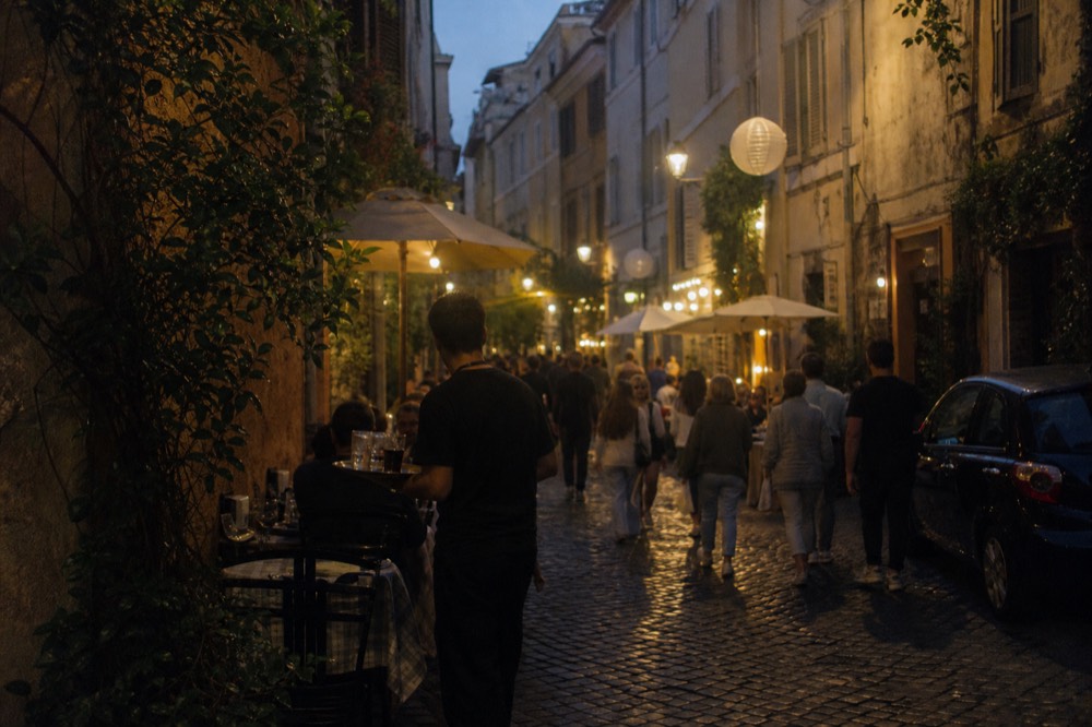 Evening crowd on a narrow Trastevere street with restaurant tables and hanging lanterns