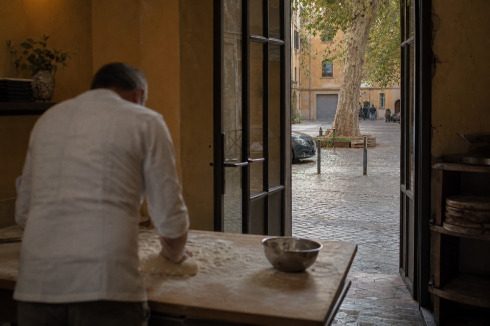 A wooden table set with fresh pasta dough, eggs, and a rolling pin in a sunlit kitchen in Rome