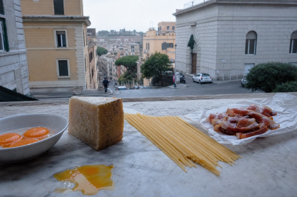Raw spaghetti, a wedge of pecorino romano cheese, a bowl of egg yolks and strips of guanciale arranged on a marble kitchen counter