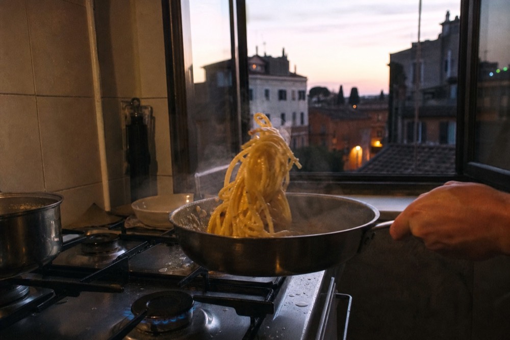 A cook's hands tossing spaghetti in a pan with a glossy egg and pecorino sauce, steam rising, in a bright home kitchen