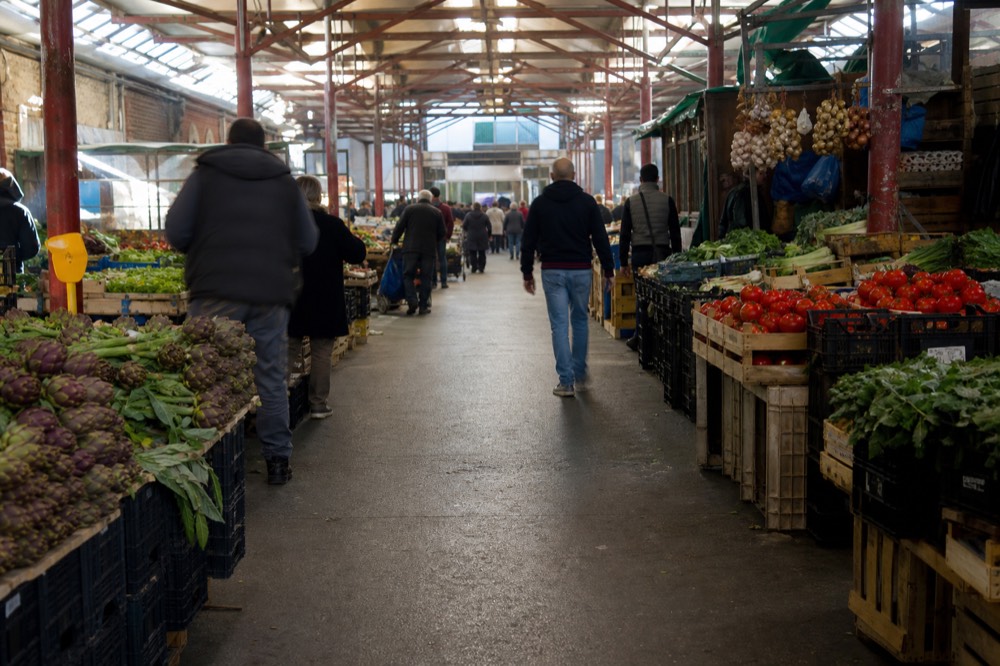 A covered market hall with produce stalls and shoppers at midday