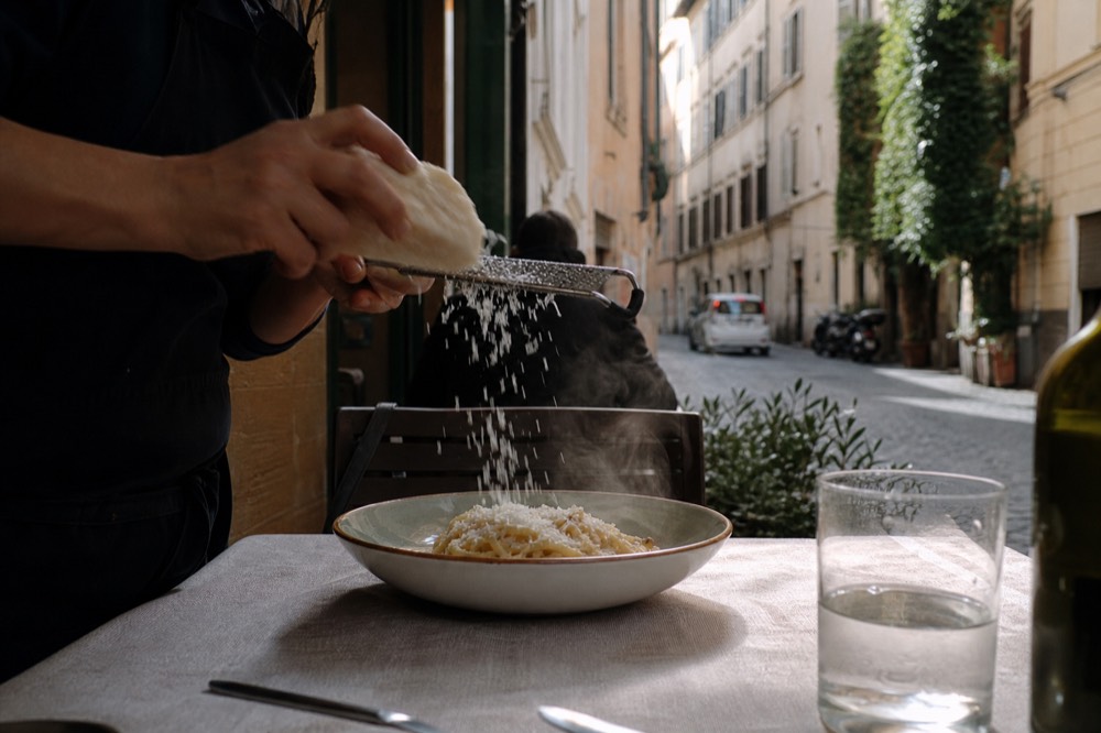 A wedge of aged pecorino romano cheese being grated over a bowl of pasta