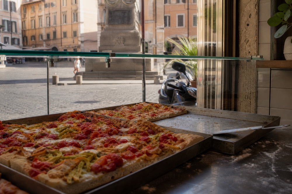 Pans of rectangular pizza al taglio with assorted toppings displayed in a bakery counter