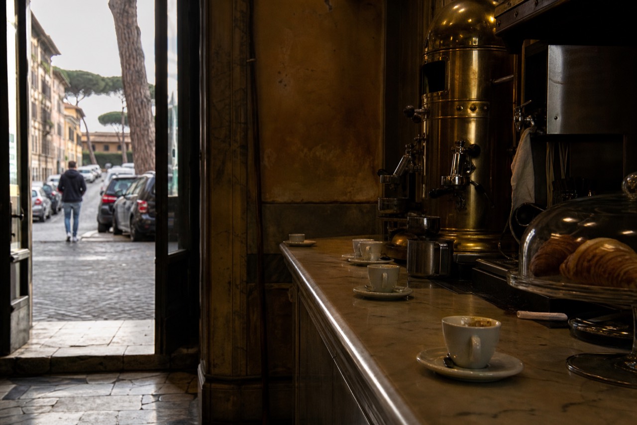 A marble bar counter in Rome with espresso cups, pastries under glass, and a brass coffee machine