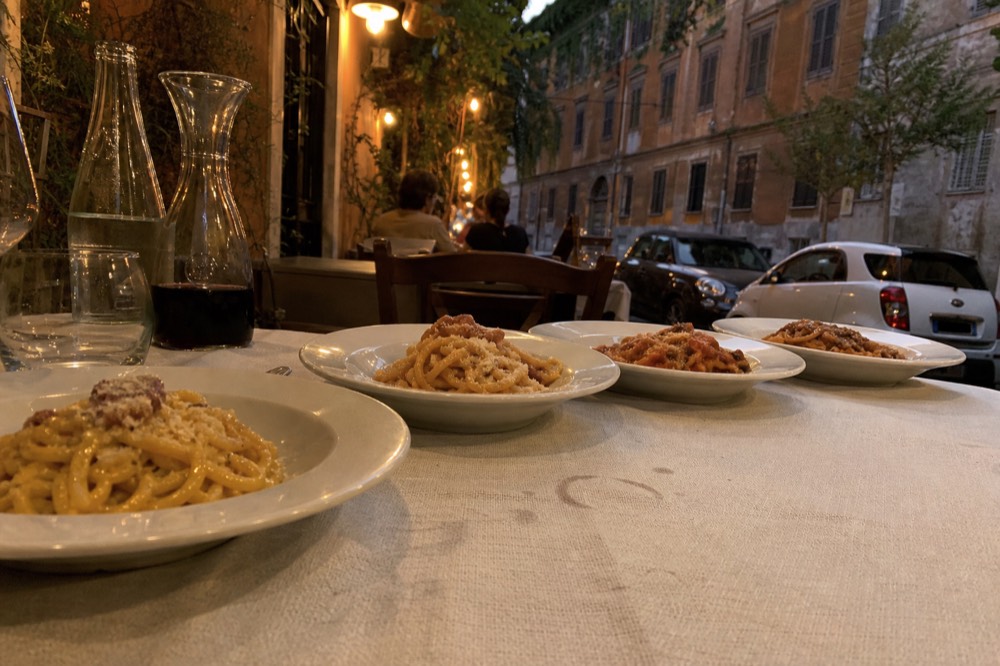 A trattoria table with four small plates of different Roman pasta dishes arranged in a row