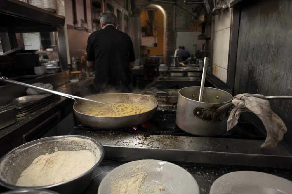 Pasta cooking in a large pan beside bowls of grated pecorino cheese in a restaurant kitchen