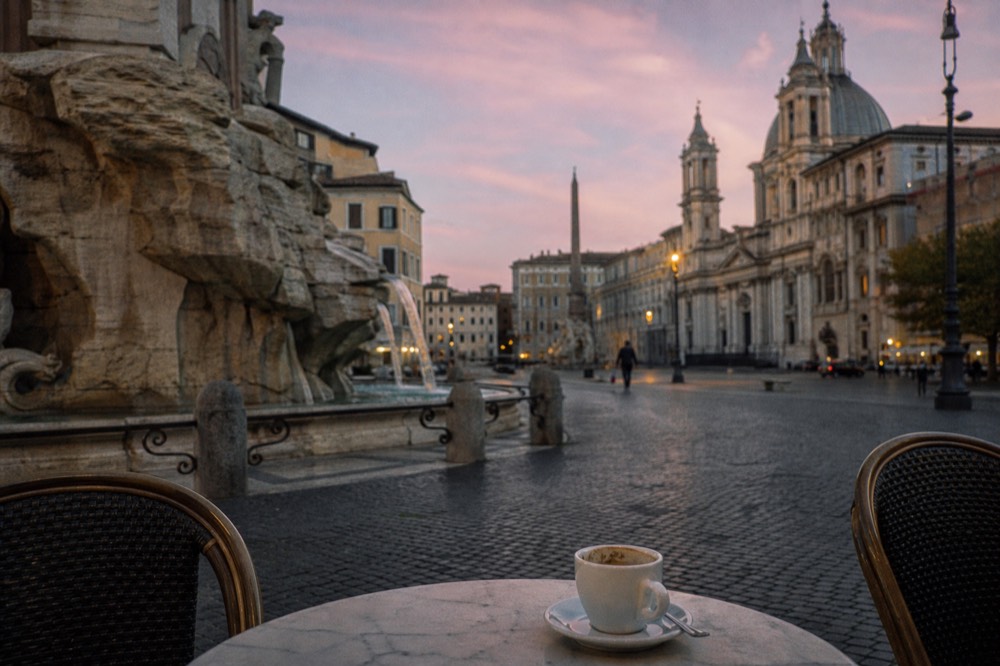 A Roman piazza at dusk with outdoor cafe tables and a fountain in the background