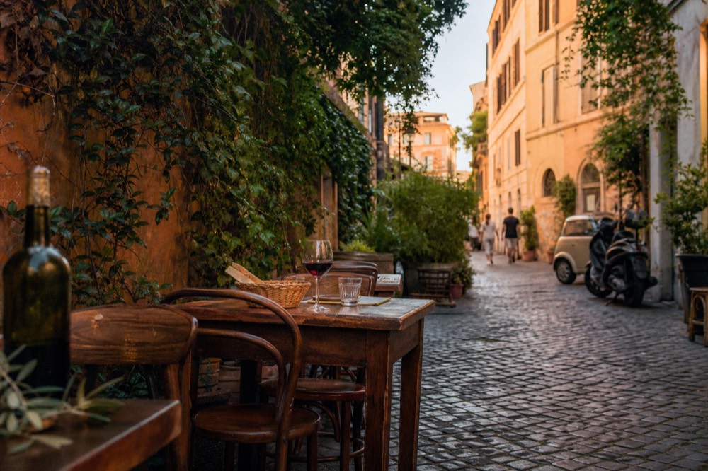 Wooden restaurant tables and chairs set out on a cobblestone lane beside ivy-covered walls