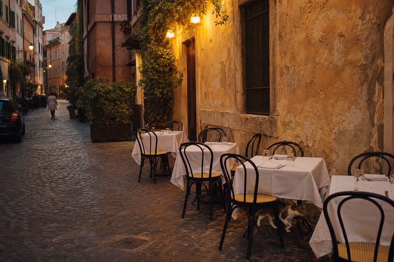 Empty trattoria tables with white tablecloths set along a cobblestone Rome street in warm evening light