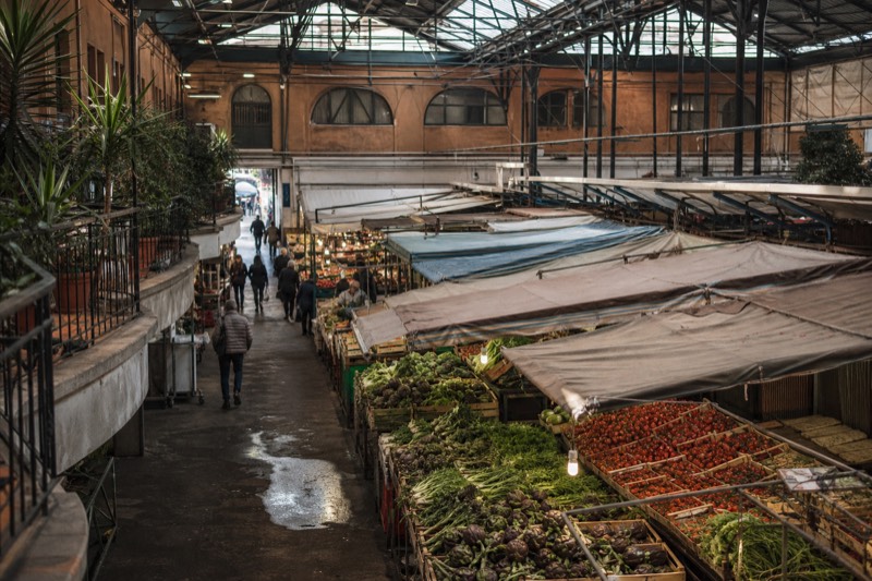 A covered food market hall in Rome, Italy, with produce stalls and shoppers between the aisles