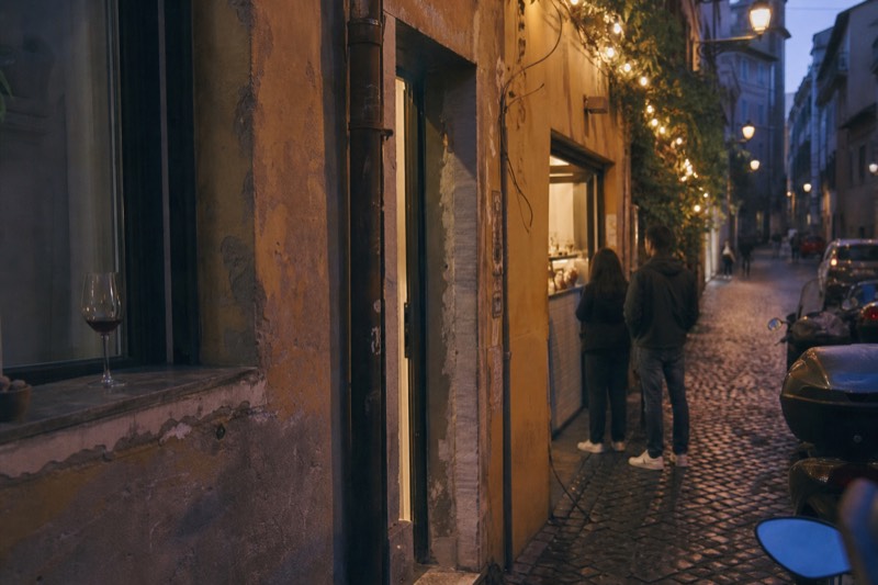 Evening food walk through a narrow Trastevere lane in Rome, with a small group gathered at a trattoria counter under warm lamplight and ivy-covered walls