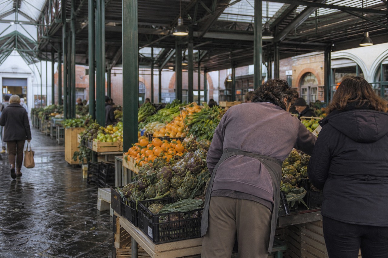 In Rome's Mercato di Testaccio, an aproned guide leans over a produce stall stacked with artichokes and oranges under a rain-flecked glass roof