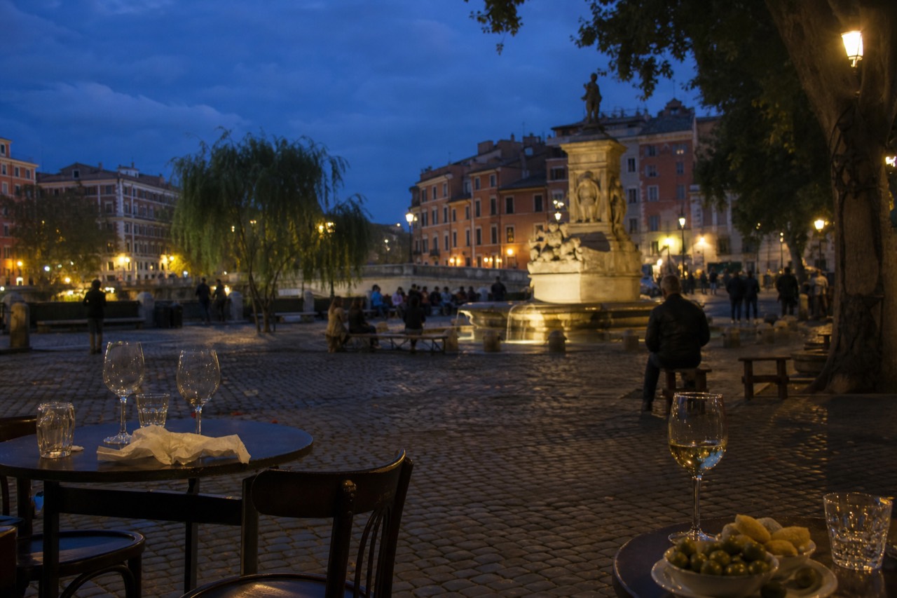 Outdoor cafe tables with wine glasses and snacks on a Rome square at dusk