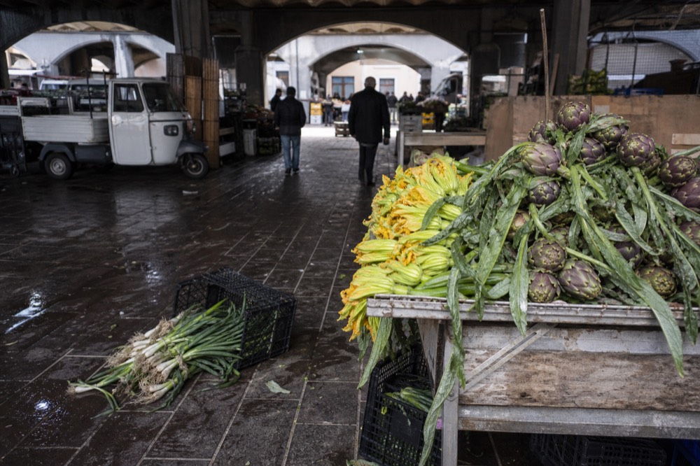 A covered Rome food market stall displaying artichokes, zucchini flowers and fresh produce
