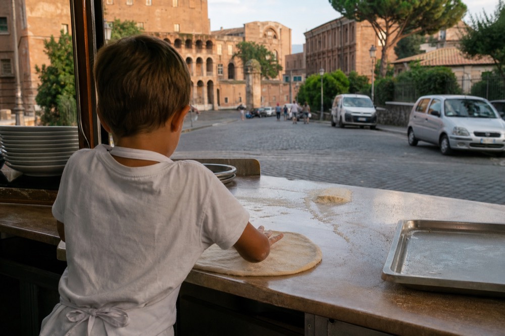 A child rolling pizza dough with flour on hands at a Rome cooking class counter