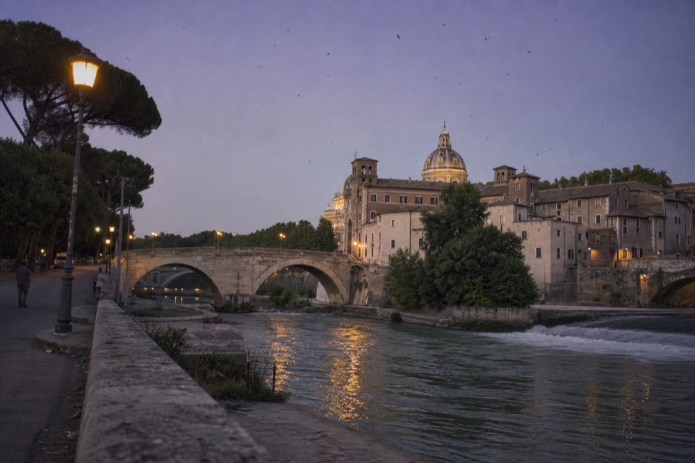 A stone bridge over the Tiber at twilight with the dome of a church lit in the distance