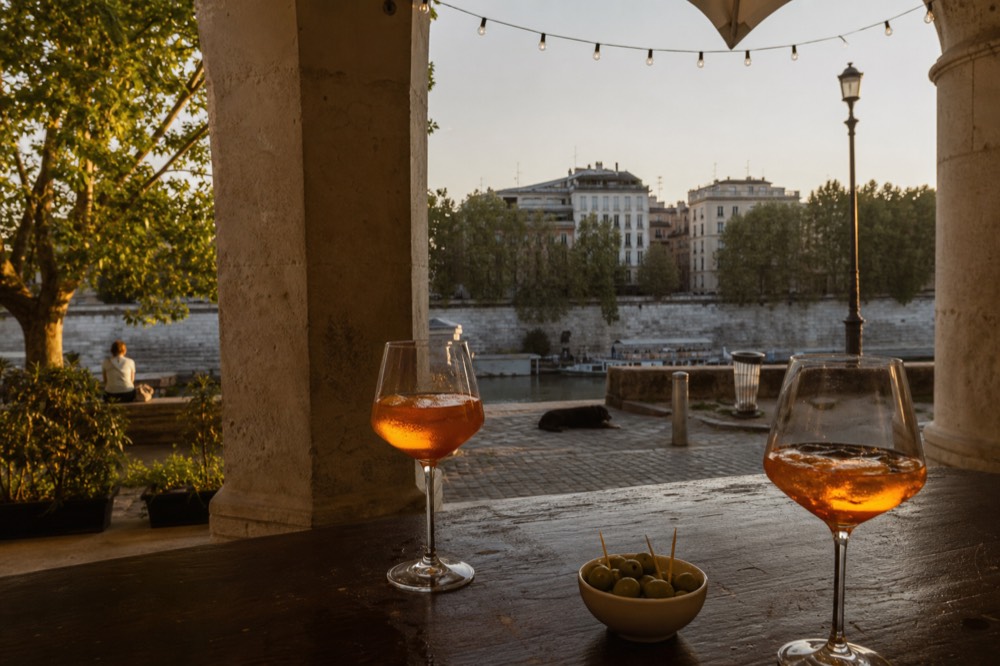 An outdoor bar counter in the early evening with glasses of amber spritz on a wooden top