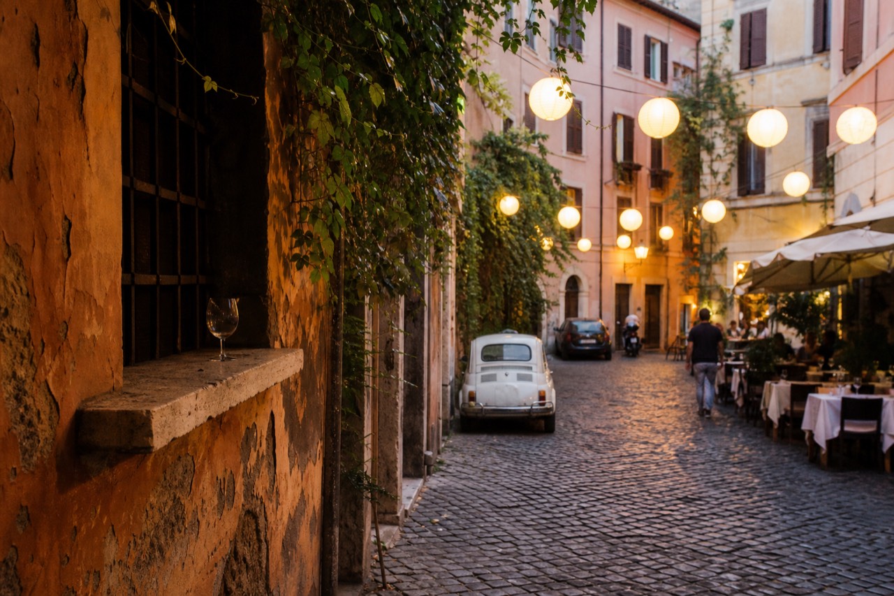 Paper lanterns glowing over a cobbled Trastevere street at dusk with restaurant tables set for dinner