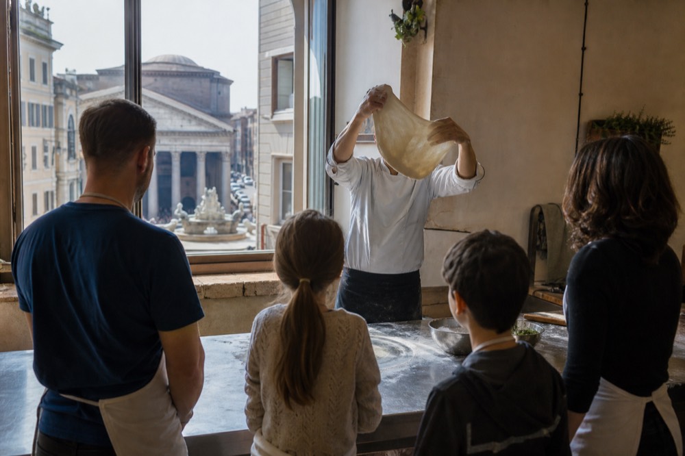 Instructor demonstrating dough stretching to a small group around a kitchen island