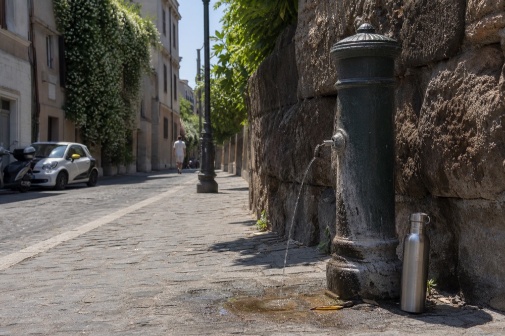 A cast-iron nasoni drinking fountain on a Rome sidewalk with water arcing from its spout