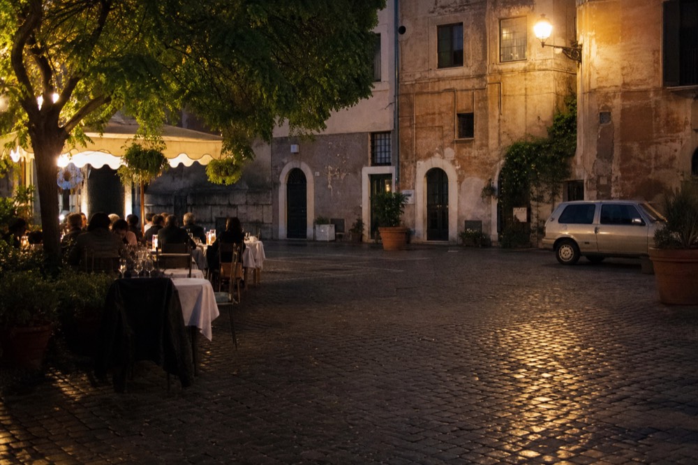 An evening piazza with lit restaurant terraces and people at outdoor tables