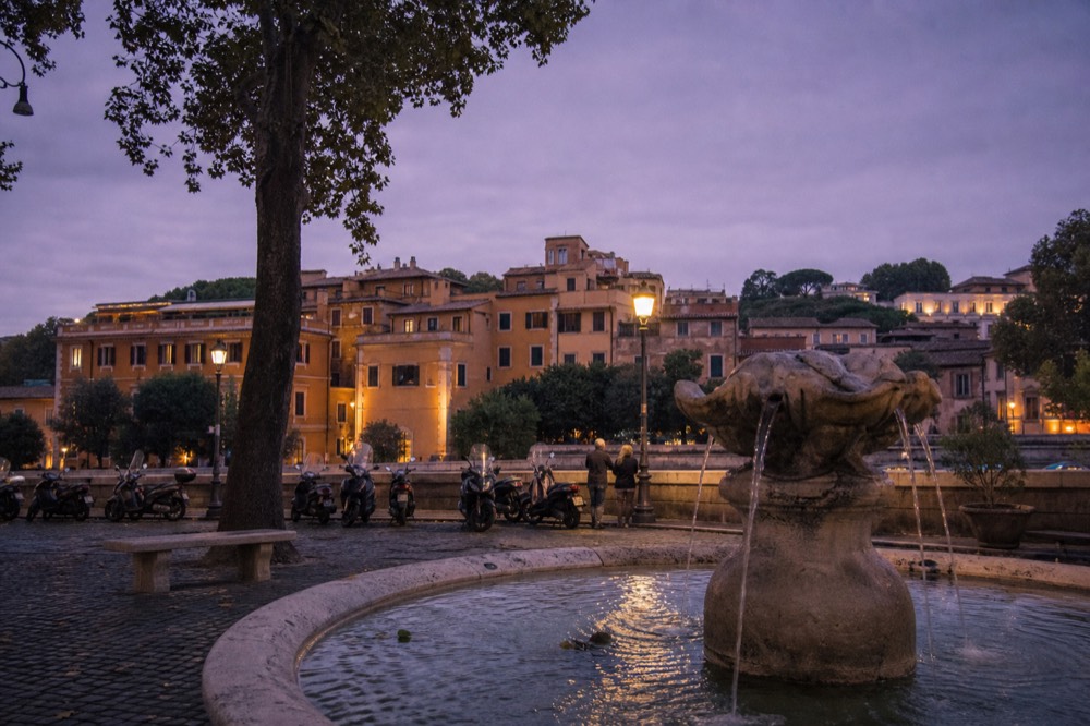 A fountain at dusk in a Roman square with parked scooters nearby