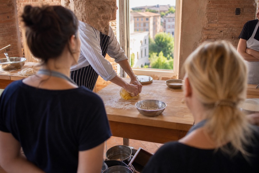 A cooking instructor demonstrating dough kneading to a small group around a kitchen island