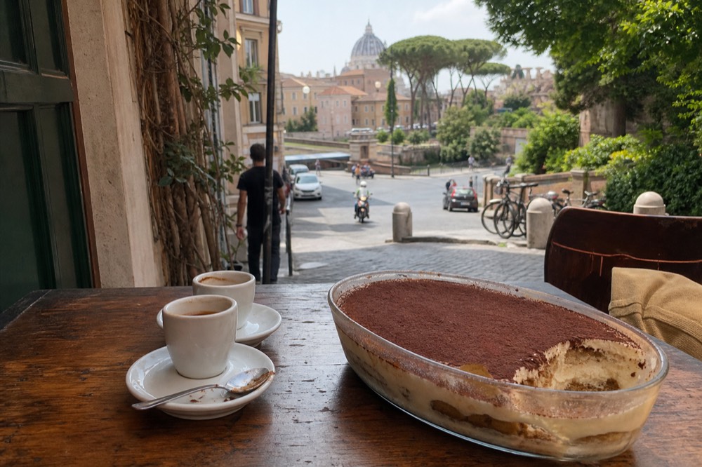 A glass dish of finished tiramisu with cocoa powder dusted on top beside espresso cups