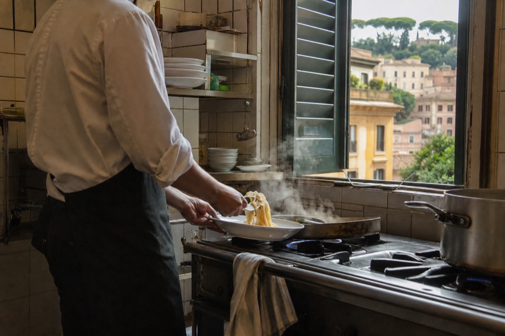 A chef plating a finished pasta dish with grated cheese in a bright Roman kitchen