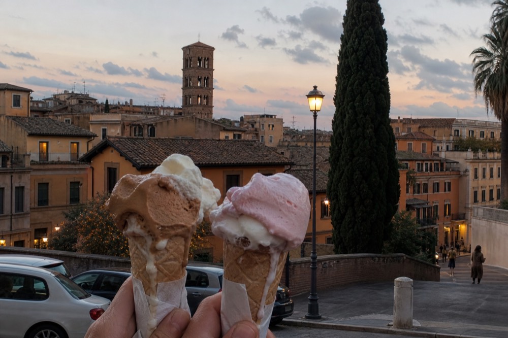 Two gelato cones held on a Rome street at dusk with historic buildings behind