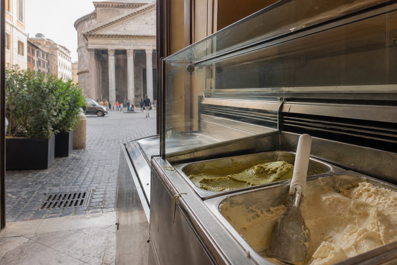 Open gelato display case with flat metal tins of pistachio and cream gelato in a Roman shop