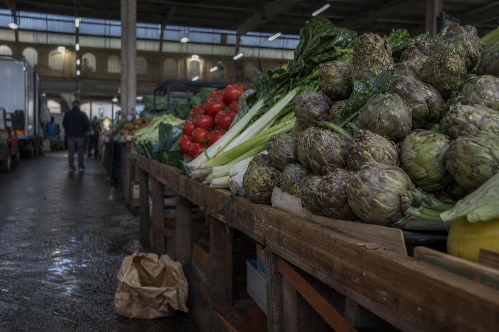 Wooden market benches loaded with artichokes, tomatoes and leafy greens beside a covered stall