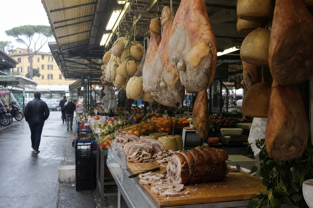 Covered market aisle with hanging cured meats and cheese wheels at a Roman market stall