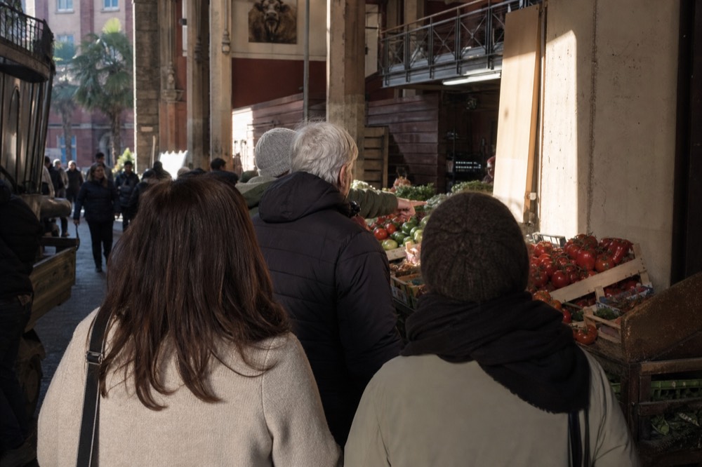 A small group of market visitors standing at a produce stall while a guide points at the produce
