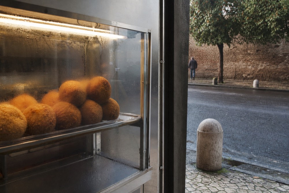 Fried rice balls stacked in a heated glass display case at a Roman snack shop