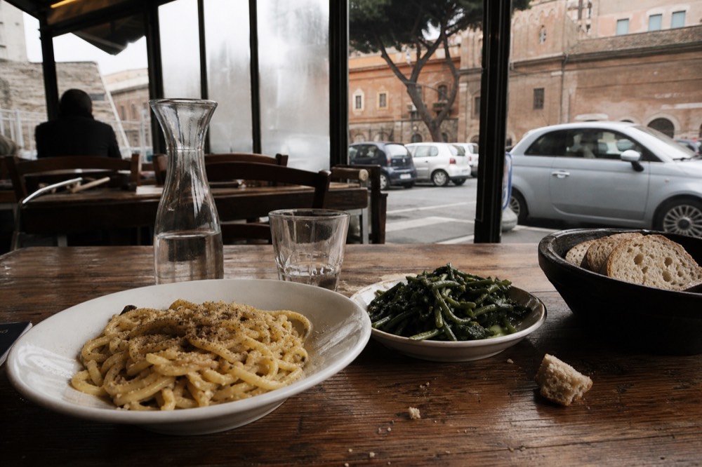 A bowl of Roman pasta with cracked black pepper and greens on a wooden table
