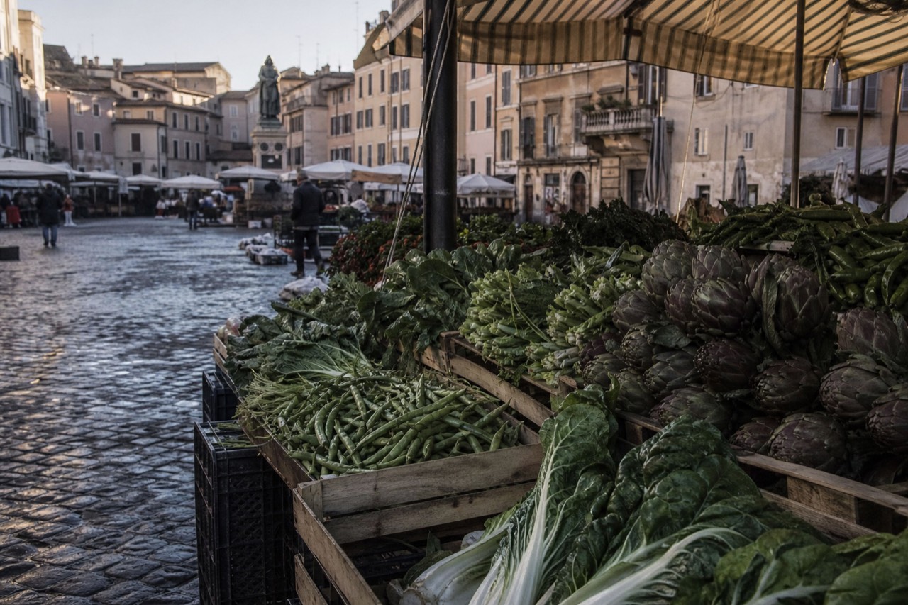 A Roman market stall piled with artichokes, broad beans and leafy greens under a canvas awning