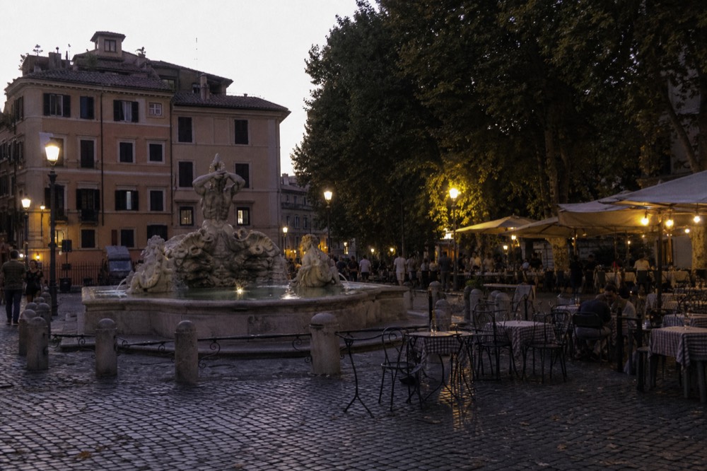 A lamplit Roman piazza at dusk with restaurant tables and people strolling