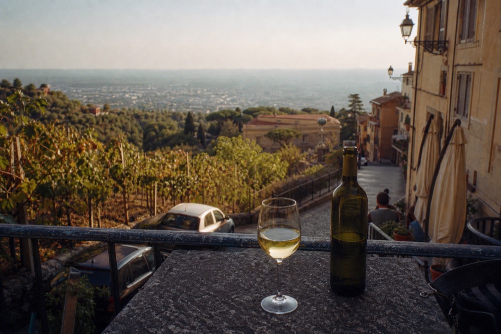 A bottle of white wine and a full glass on a table overlooking hillside vineyards
