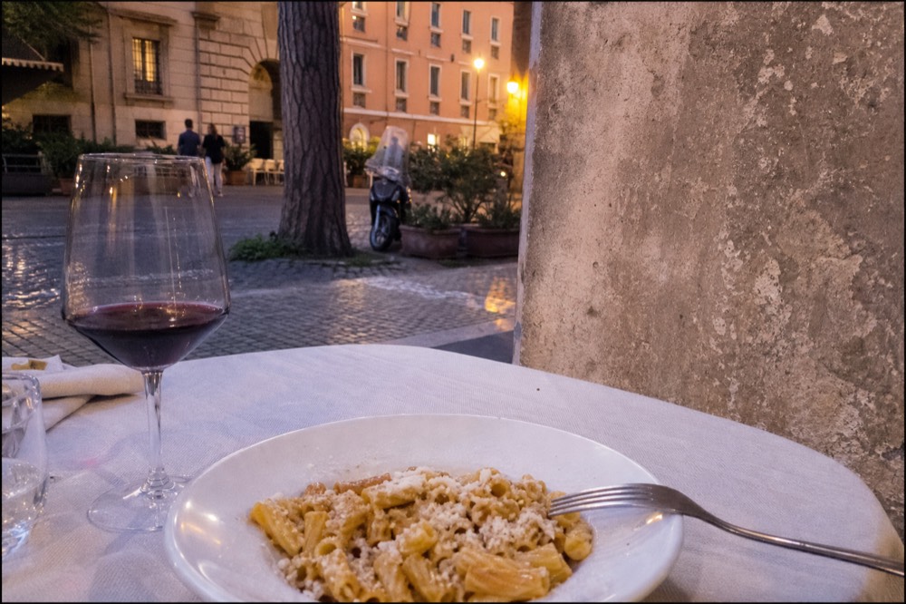 A glass of red wine beside a plate of pasta with pecorino cheese at a restaurant table